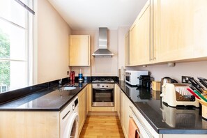 A compact kitchen with a stove, microwave, and sink, wooden cabinets, and a window. - Charming Two-Bed in South Kensington (London)