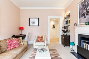Ein Wohnzimmer mit Sofa, Sessel, Couchtisch, Kamin, Bücherregal und gerahmten Kunstwerken.