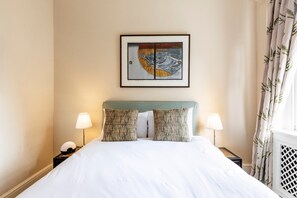 A hotel room with a bed, two bedside lamps, a framed artwork above the bed, and a window with curtains. - Charming Two-Bed in South Kensington (London)
