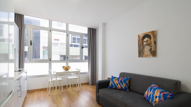 A modern living room with a grey sofa, two patterned cushions, a white table with chairs, and a window with curtains. - Beachfront Apartment in Las Palmas by Las Canteras (Las Palmas de Gran Canaria)