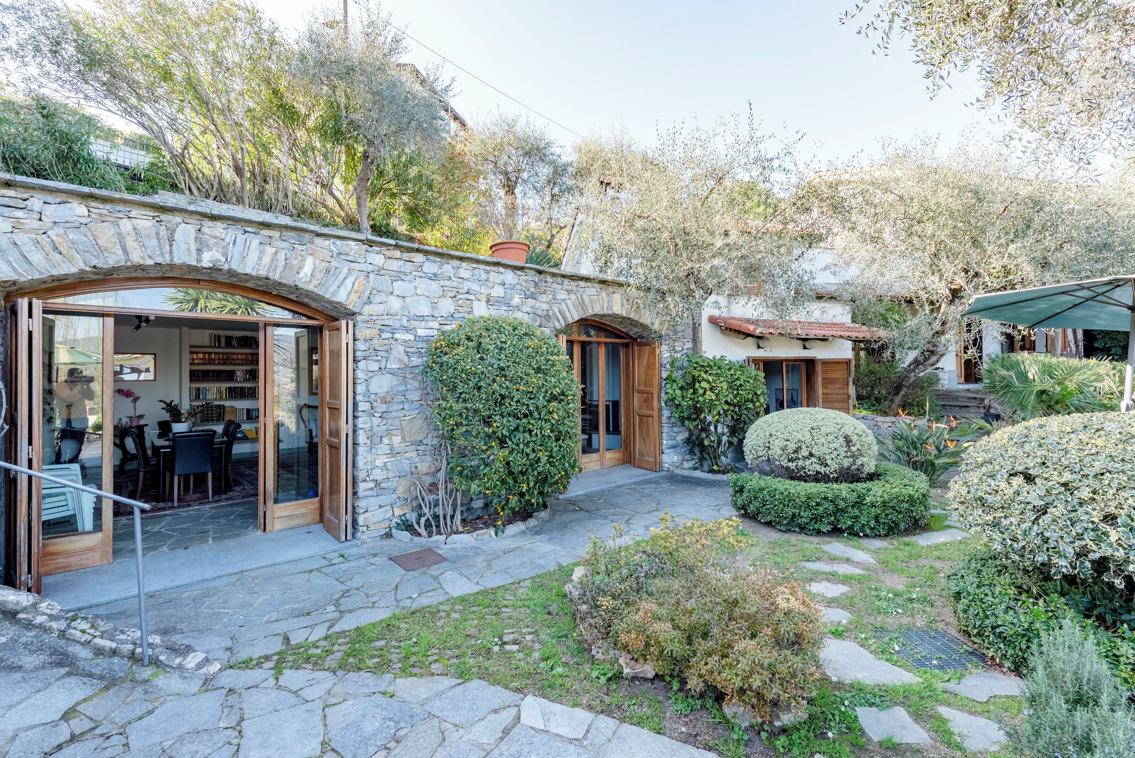 Edificio de piedra con entrada de puerta de cristal, un patio con suelo de piedra y un jardín con diversas plantas y un pequeño árbol.