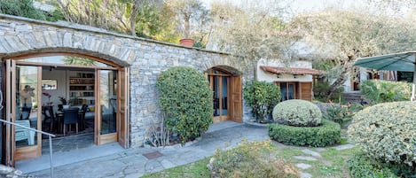 A stone building with a glass door entrance, a patio area with stone flooring, and a garden with various plants and a small tree.