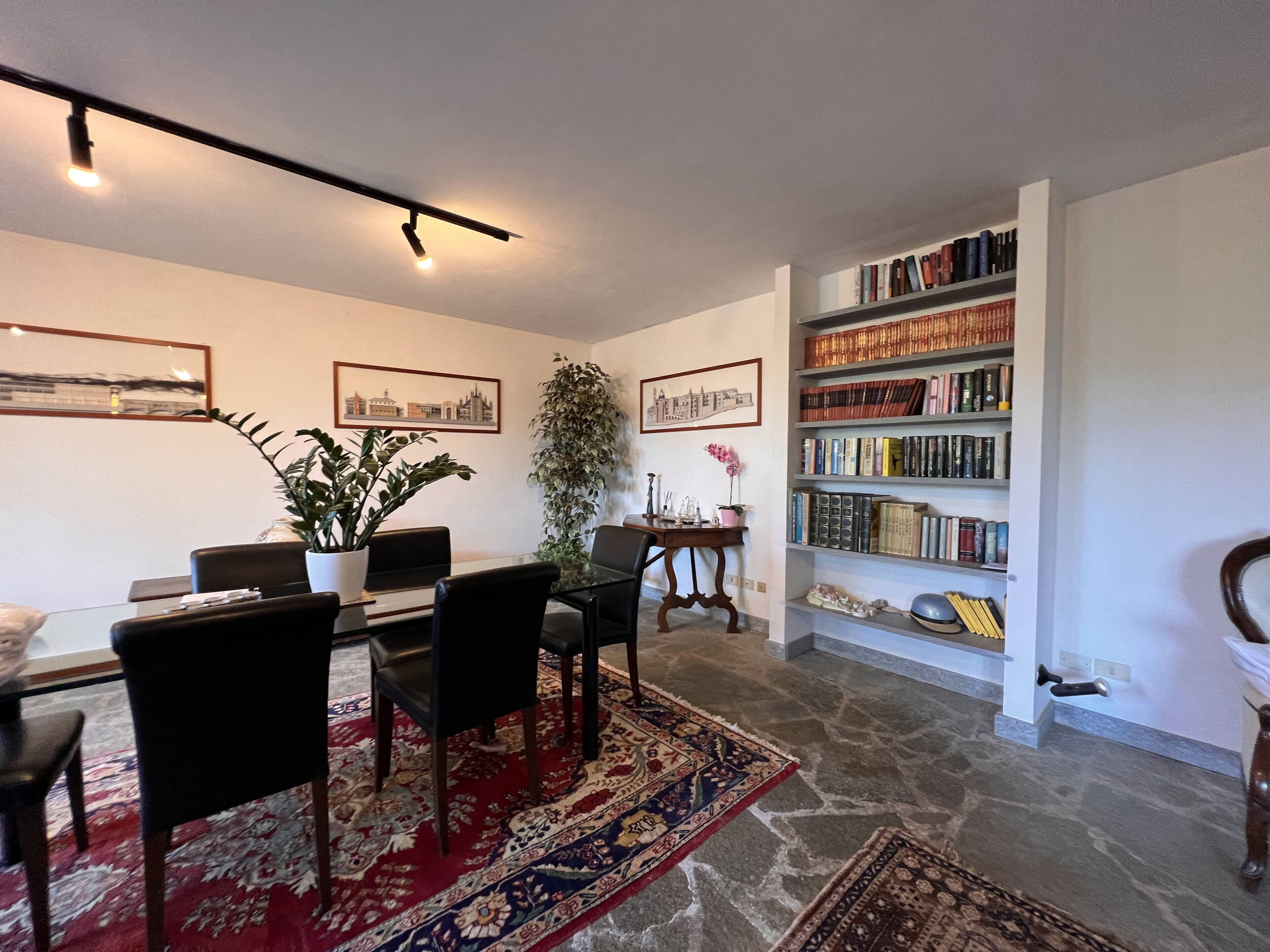 Ein Essbereich mit Tisch, Stühlen, einem roten Teppich mit Muster, einem Bücherregal voller Bücher und gerahmten Bildern an den Wänden.