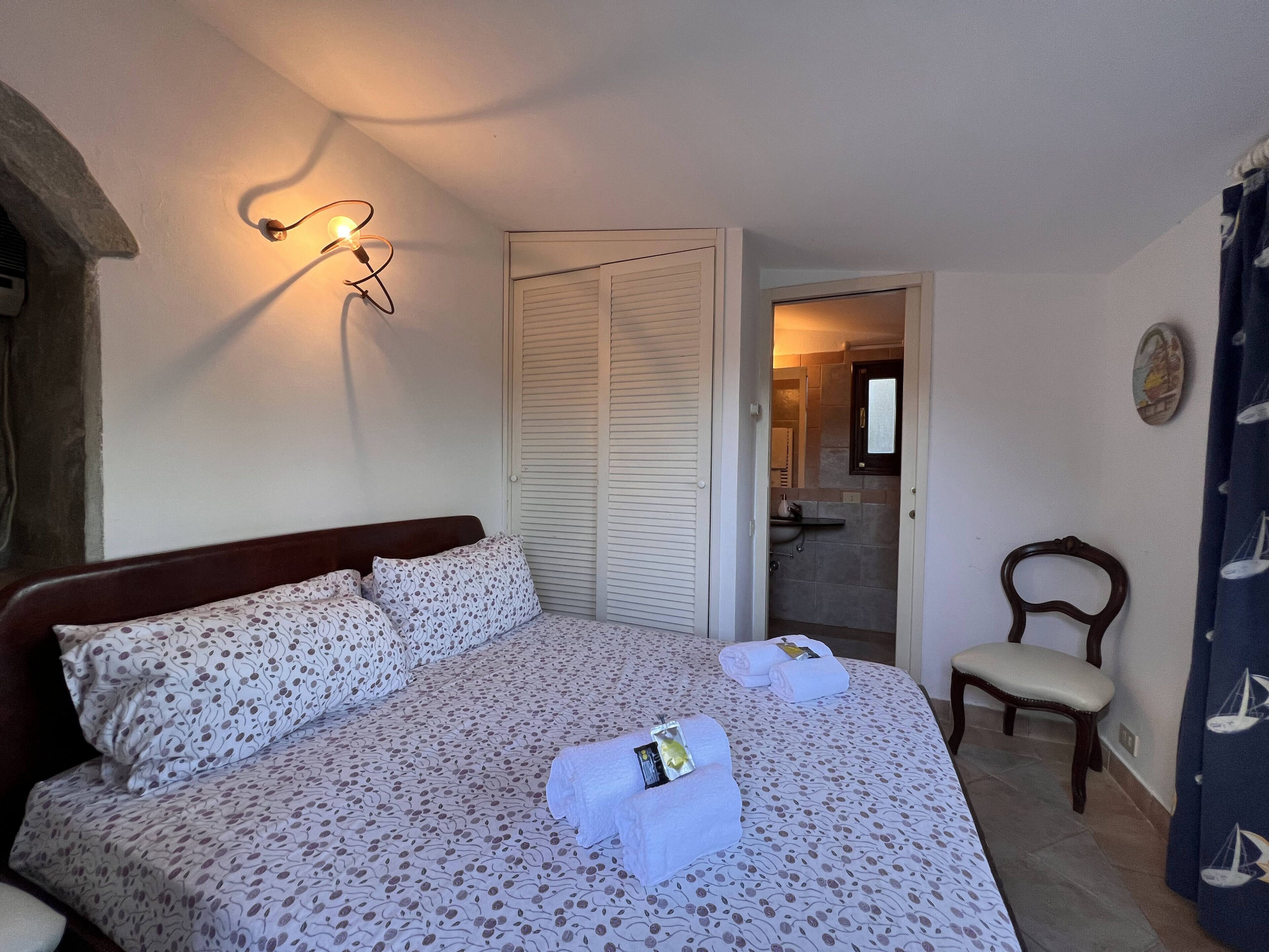 A neatly made bed with patterned bedding, a bedside lamp, a chair, and a bathroom visible through an open door.