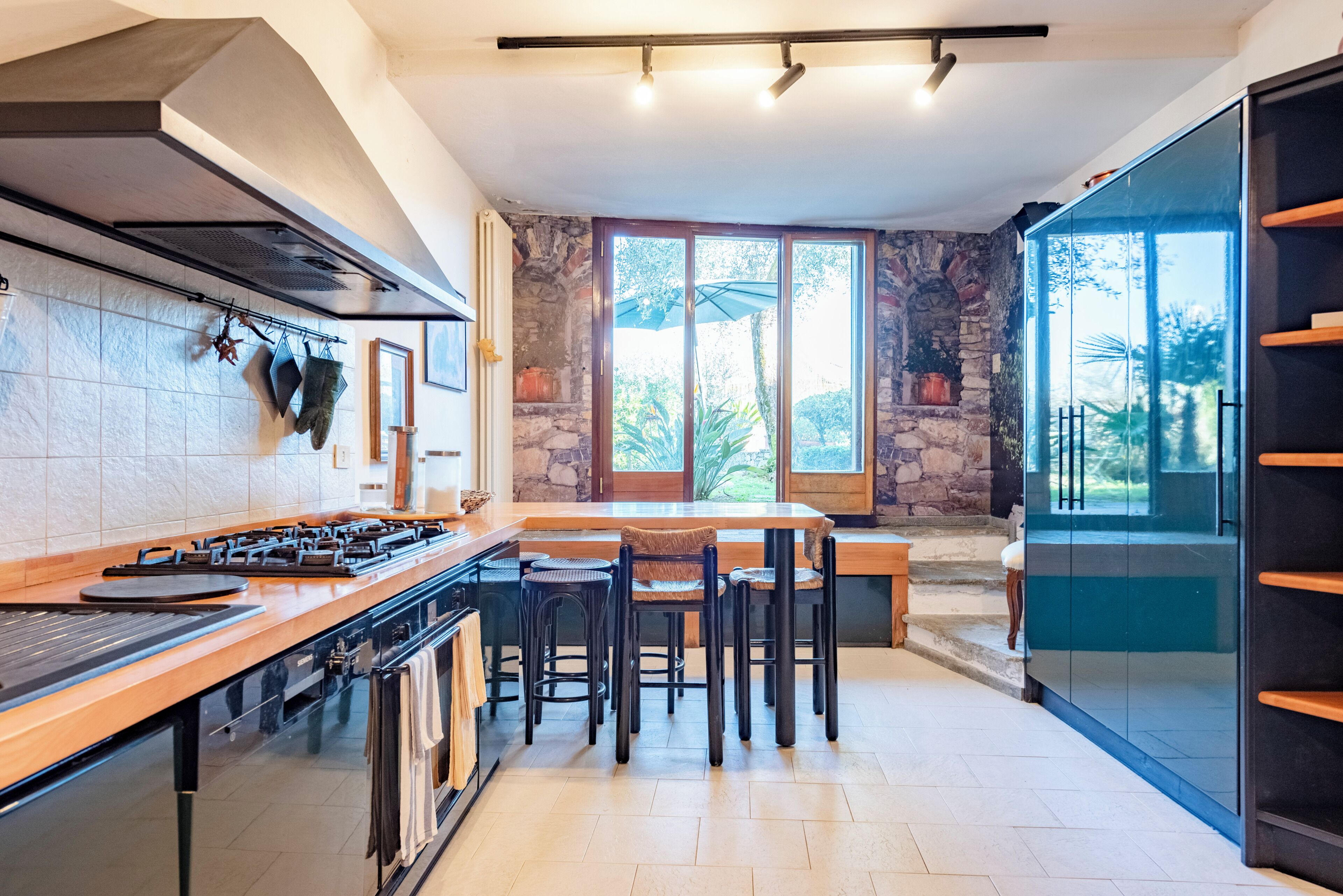 Una cocina moderna con estufa a gas, taburetes de bar, un muro decorativo de piedra y ventanas amplias.
