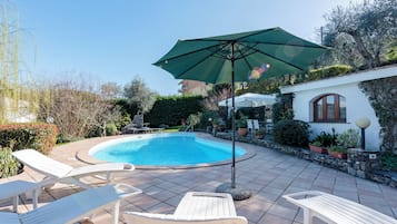 Une piscine entourée de verdure et d’un mur de pierre, avec des chaises longues et un parasol.