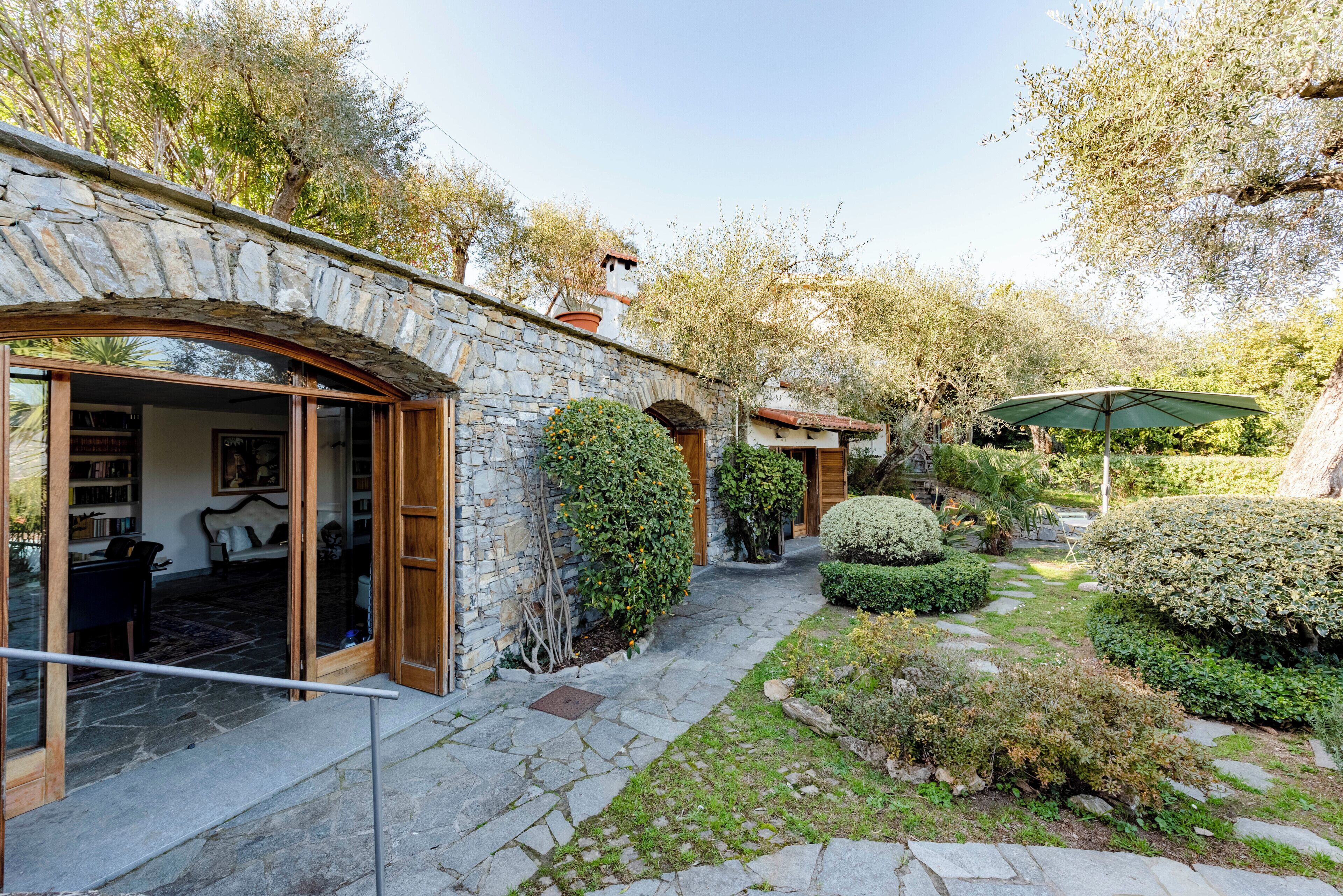 A stone-walled building with a glass door entrance, a stone pathway, and a well-maintained garden with various shrubs and a palm tree.