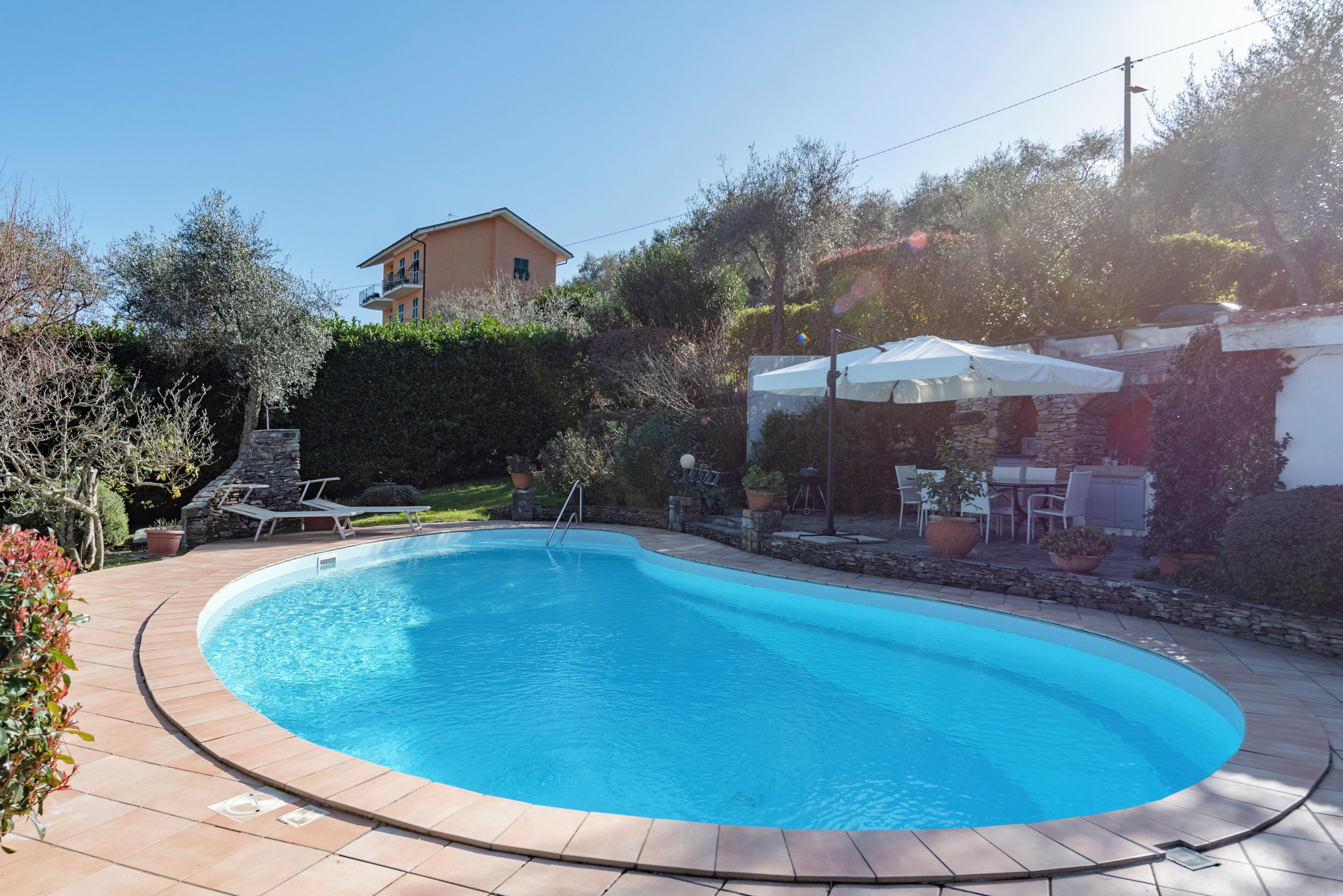 A swimming pool with a stone border, a patio area with outdoor furniture, and a building in the background.