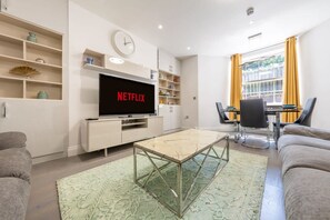 A modern living room with a flat-screen TV displaying 'NETFLIX,' a dining area with a table and chairs, and a sofa. - Super Deluxe 3 Bedroom Apartment (London)