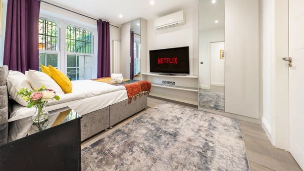 Ein modernes Schlafzimmer mit Bett, Fernseher, Spiegel und einem Fenster mit Vorhängen.