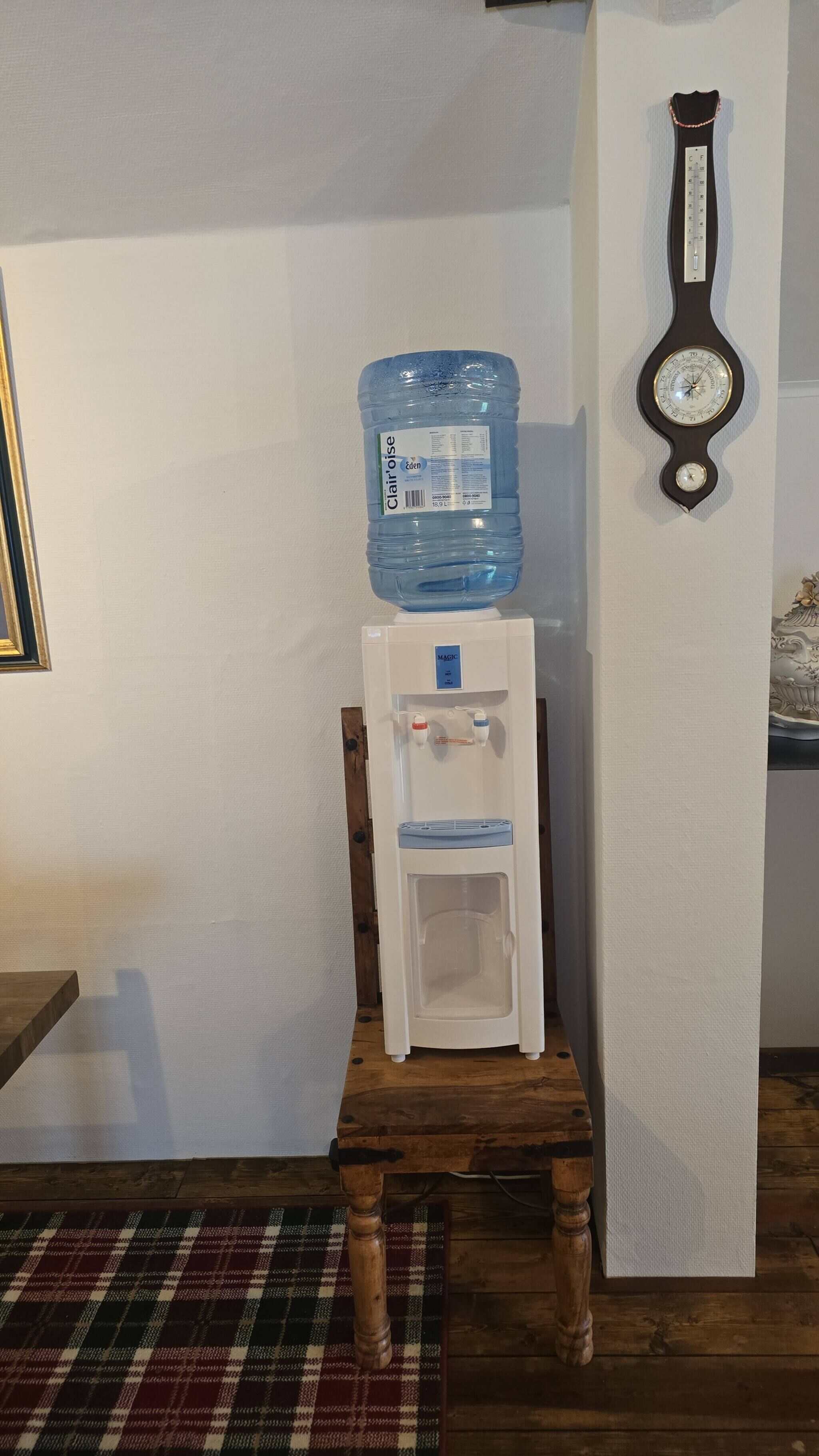 Un dispensador de agua sobre un taburete de madera, una jarra de agua y un reloj fijado a la pared.