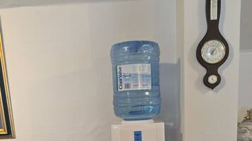 Un dispensador de agua sobre un taburete de madera, una jarra de agua y un reloj fijado a la pared.