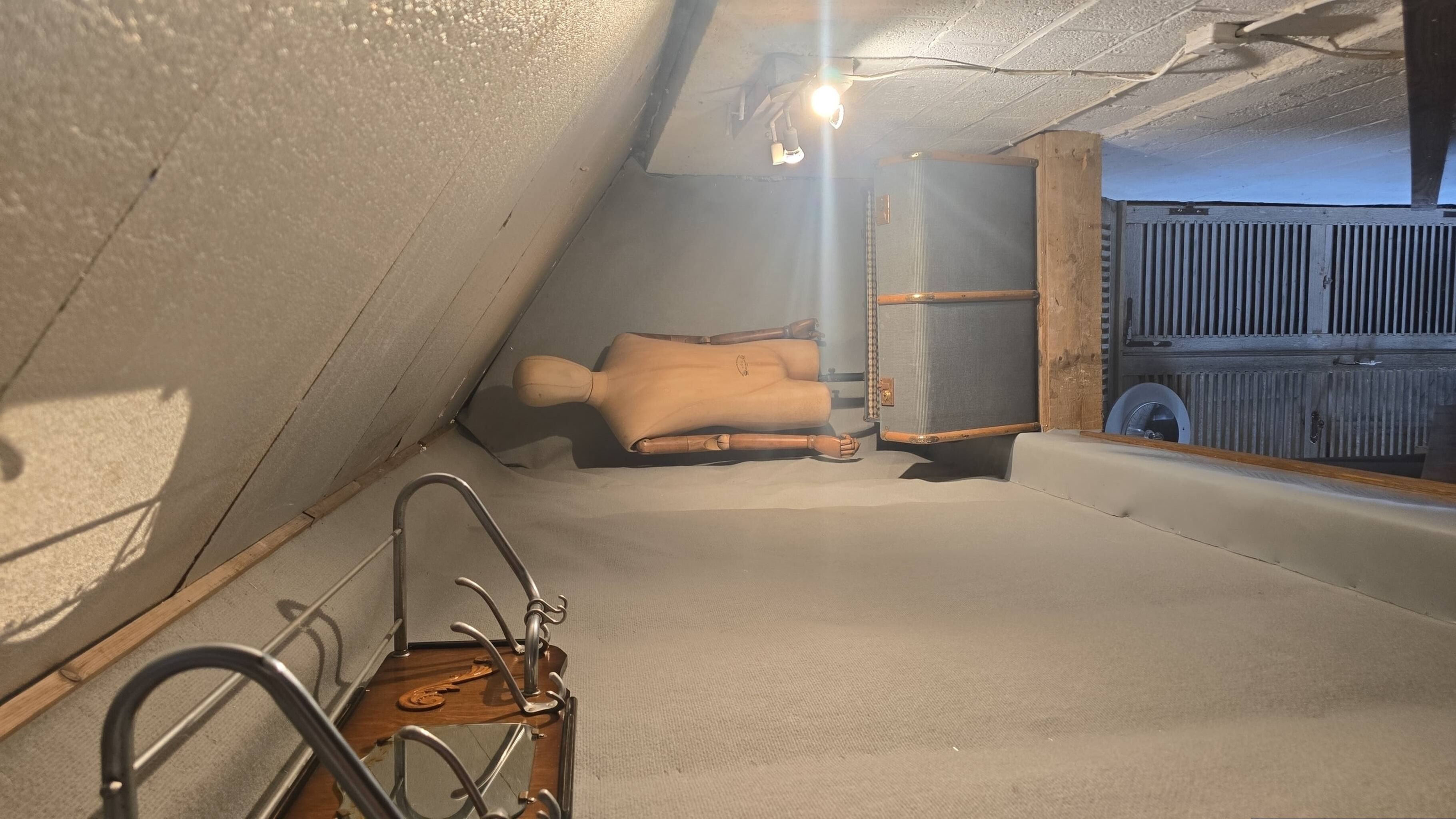 A room with a sloped ceiling, a wooden chair, a metal chair, and a suitcase.
