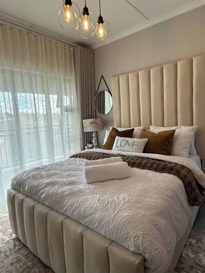 A modern bedroom with a large bed, a headboard, a bedside table with a lamp, and a view of the city through large windows. - Waterfall Executive suite (Midrand)