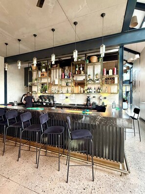A bar area with a counter, bar stools, hanging pendant lights, and shelves with bottles and decorative items. - Waterfall Executive suite (Midrand)