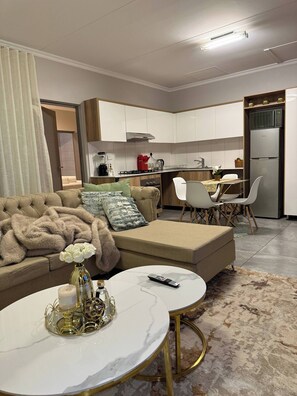 A modern living room with a sofa, ottoman, and a round coffee table with a vase and candle. A kitchen with white cabinets and a refrigerator is visible in the background. - Waterfall Executive suite (Midrand)