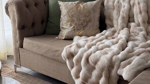Un canapé beige avec un dossier capitonné, un coussin vert, un coussin décoratif à motifs dorés et un plaid en fausse fourrure blanche.