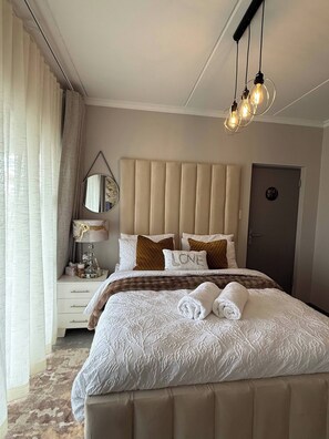 A hotel room with a large bed, a bedside table with a lamp, a mirror, and a door. - Waterfall Executive suite (Midrand)