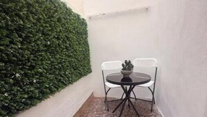 Un petit espace extérieur clos, avec une table ronde et deux chaises, entouré d’un mur blanc et d’une haie verte.
