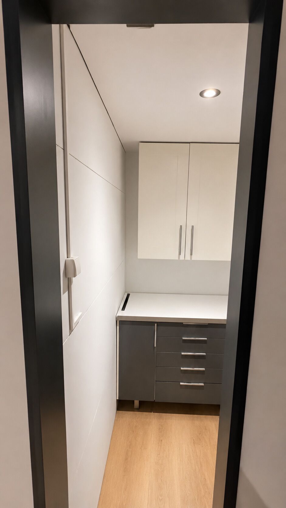 Un couloir étroit mène à une pièce où se trouvent un bureau, des tiroirs et un luminaire au plafond.