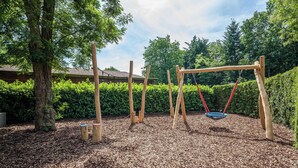 Une aire de jeux avec des balançoires et un toboggan, entourée d’arbres et recouverte de paillis.