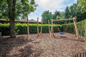 Un parque infantil con columpios y un tobogán, rodeado de árboles y acolchado de mulch.