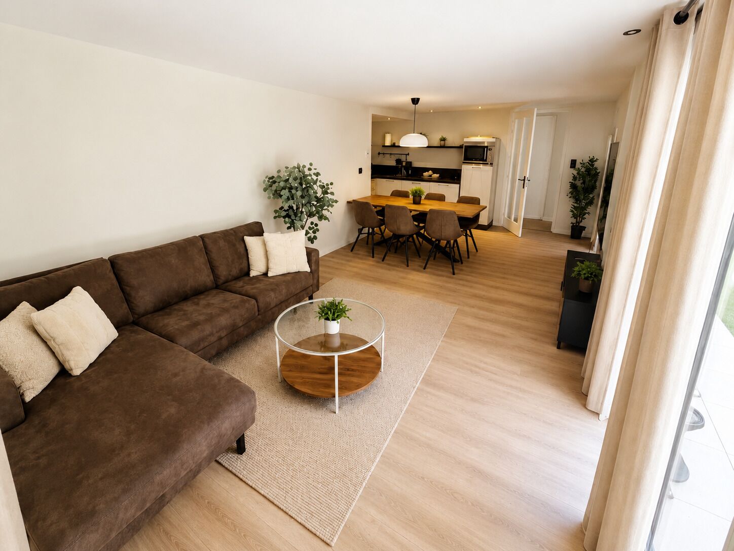 Una sala de estar moderna con un sofá marrón, una mesa de centro de vidrio y un comedor con una mesa y sillas de madera.