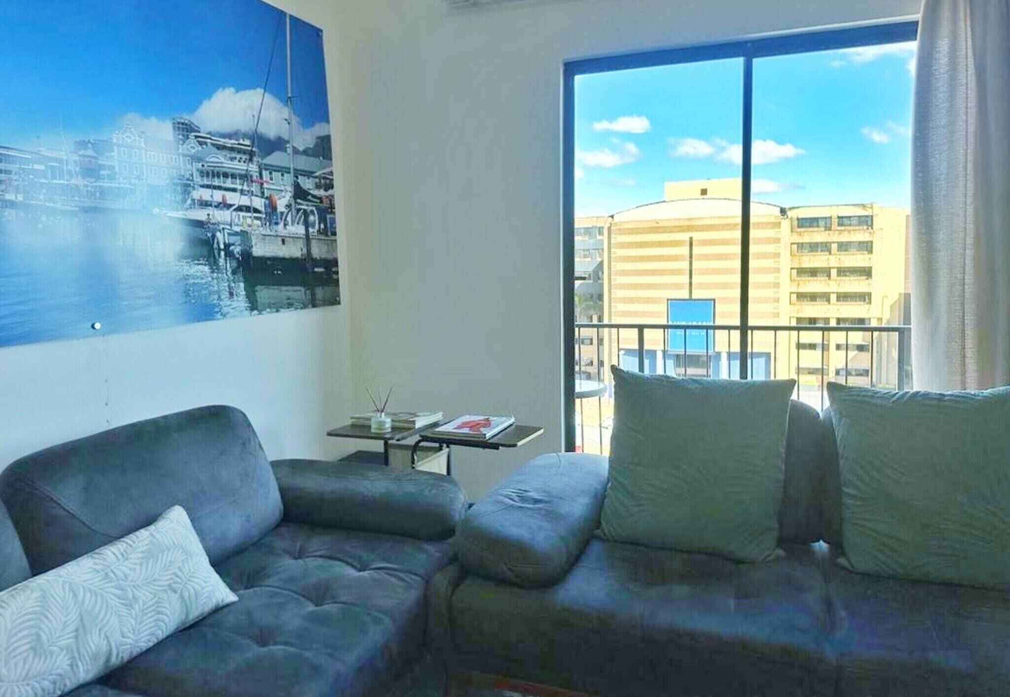 Una sala de estar con un sofá, una mesita con un libro y un ventanal con vistas a edificios.
