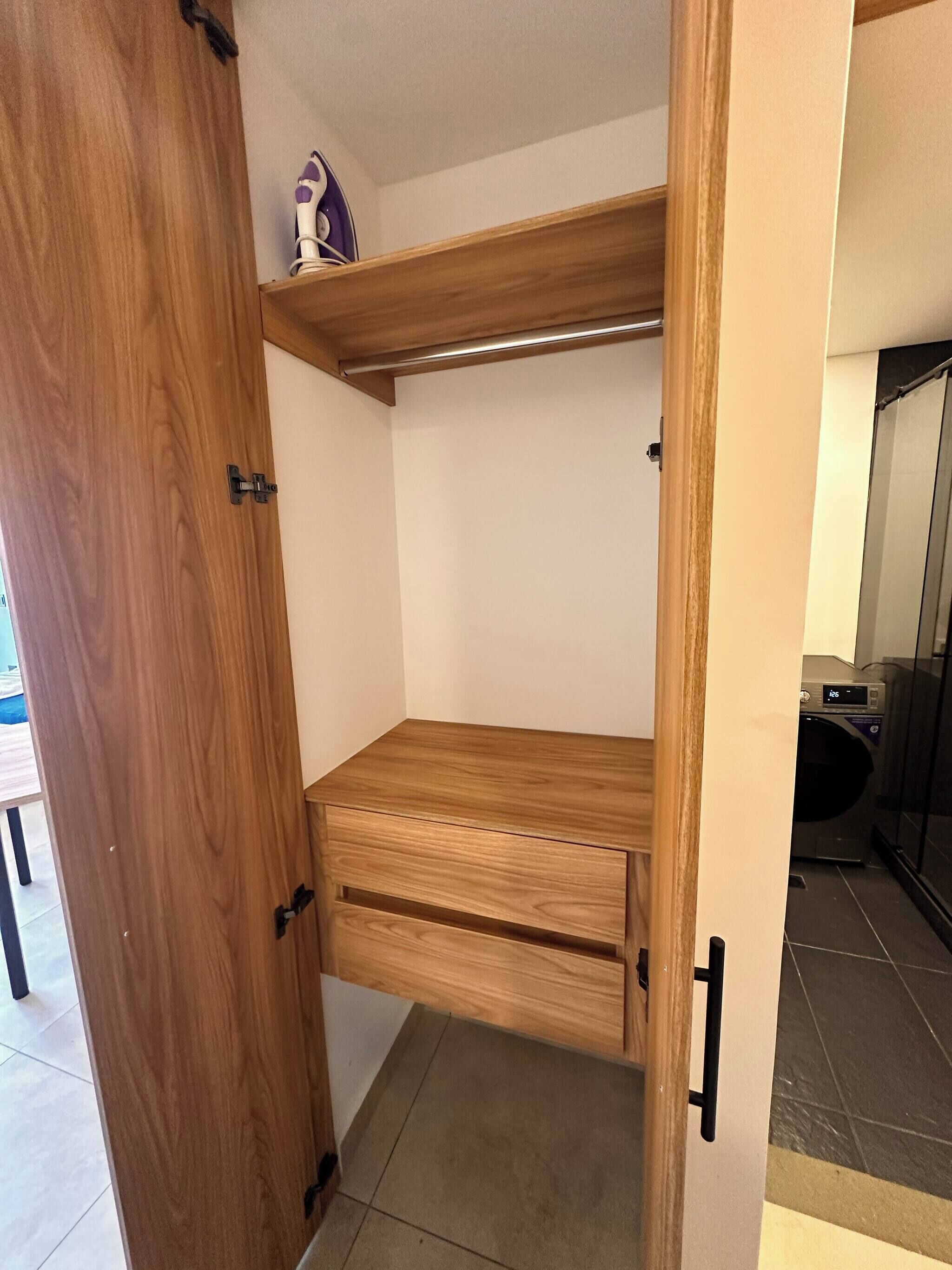 Une armoire en bois avec une étagère et une tringle à vêtements, un petit objet blanc sur l’étagère, et un lave-linge à l’arrière-plan.