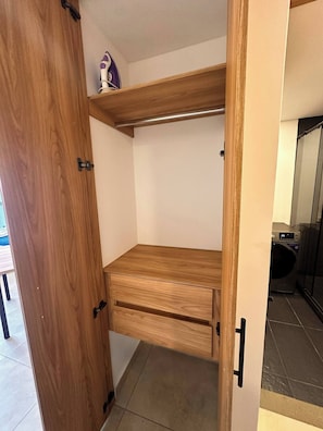 Ein Holzschrank mit Ablagefläche und Kleiderstange, ein kleines weißes Objekt auf der Ablagefläche sowie eine Waschmaschine im Hintergrund.