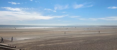 Una playa extensa con gente caminando, cielo despejado y montañas a lo lejos.