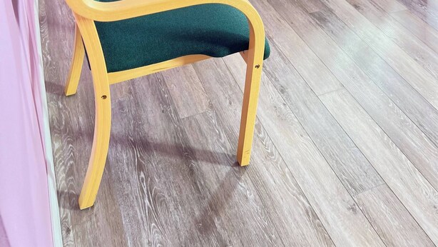 Une chambre avec un lit à cadre en bois, un fauteuil recouvert d’un tissu vert, une table de chevet blanche avec une lampe, et un rideau bleu.