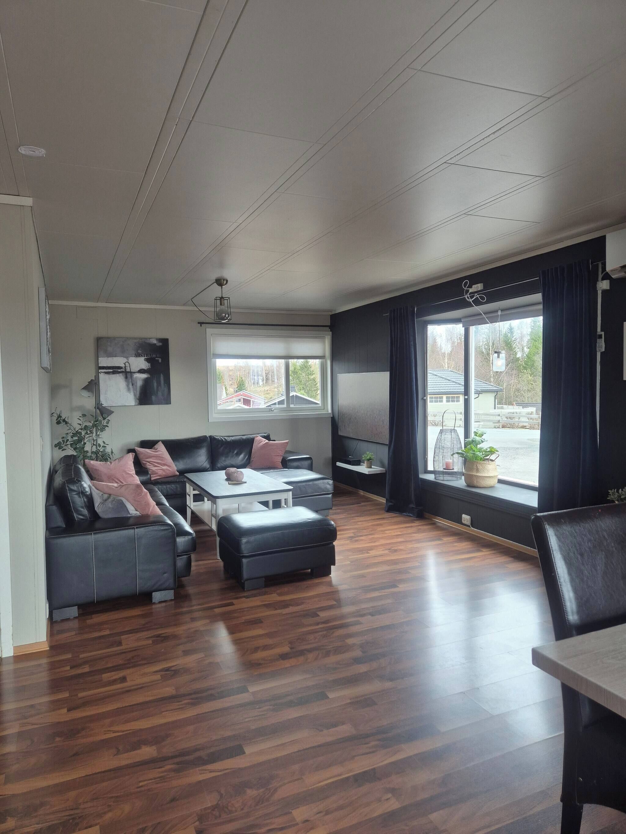 A modern living room with a sectional sofa, ottoman, and a dining table. There are two windows with curtains, a ceiling light, and a wall-mounted TV.