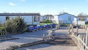 A caravan park with multiple caravans parked in rows, a gravel parking area, and a wooden bridge leading to a fenced area. - MKRJ LUXURY PLUS – Sleeps 8 - Pet Friendly - LARGE En-suite- mp619 (Camber)