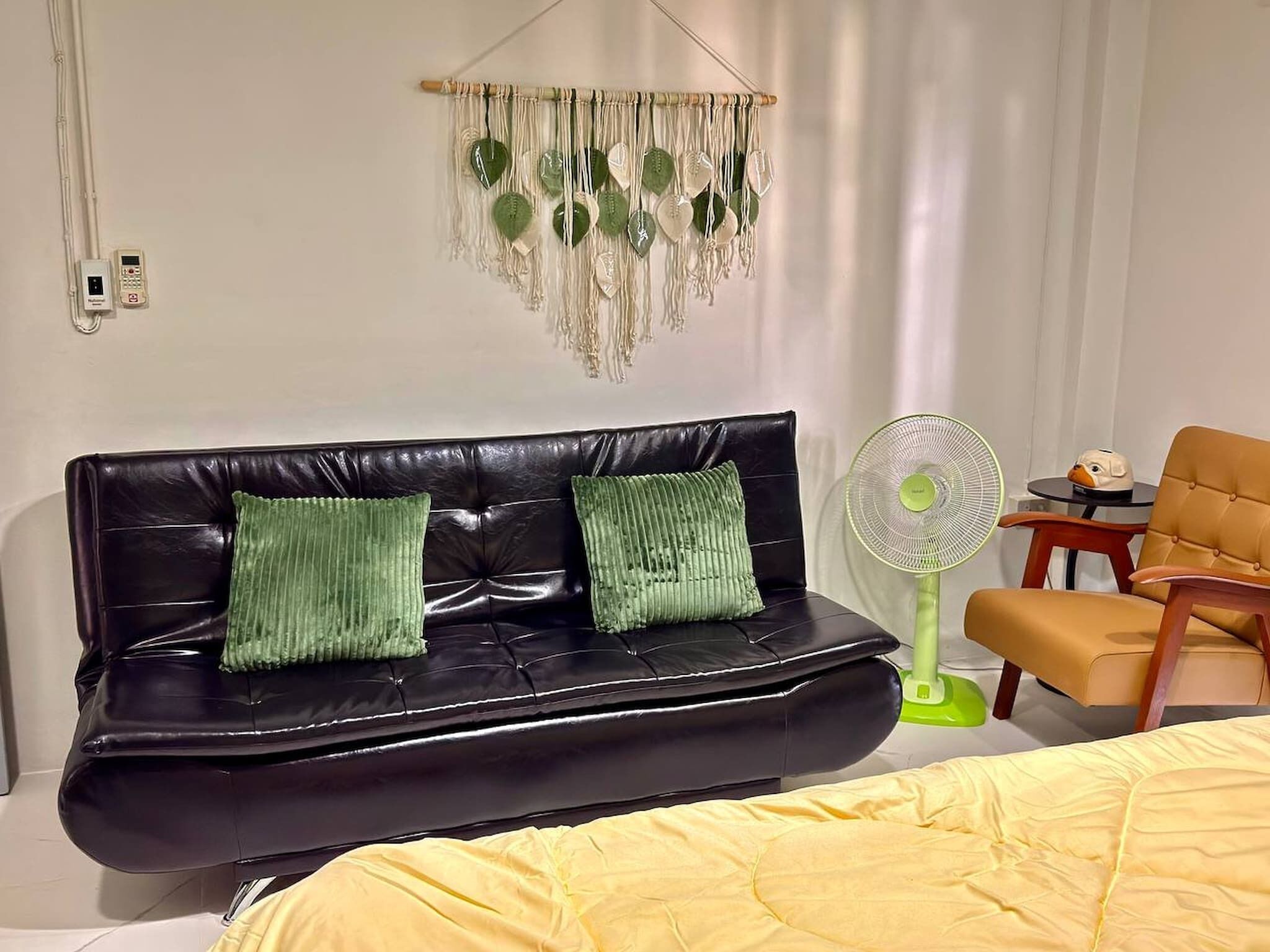 A room with a black leather sofa, two green cushions, a yellow bedspread, a green standing fan, and a small table with a decorative item.