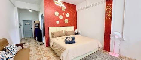 A hotel room with a bed, a chair, a desk, a wall with decorative items, and a ceiling fan.