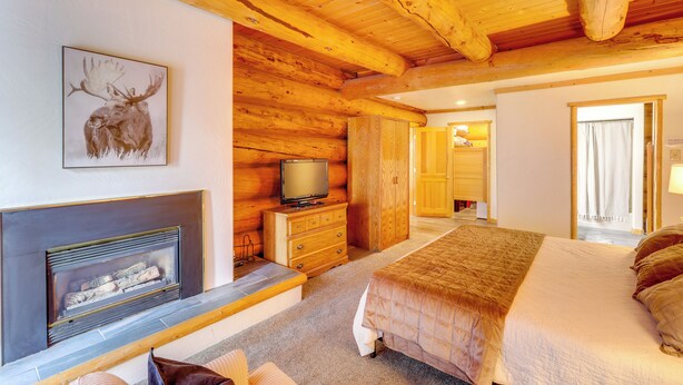 Interior de uma cabana de madeira com lareira, uma cama, uma televisão e teto de madeira.