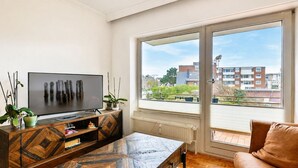 Una sala de estar moderna con televisión, mesa de centro de madera, sofá y ventanas amplias con vistas a edificios.