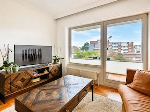 Ein modernes Wohnzimmer mit Fernseher, Couchtisch aus Holz, Sofa und großen Fenstern mit Blick auf Gebäude.