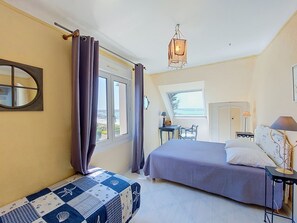 A bedroom with a bed, a bench, a table, a lamp, a mirror, and a view of the sea.