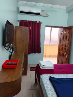 A bedroom with a bed, a wooden desk with a TV, an air conditioner, and a red basket.