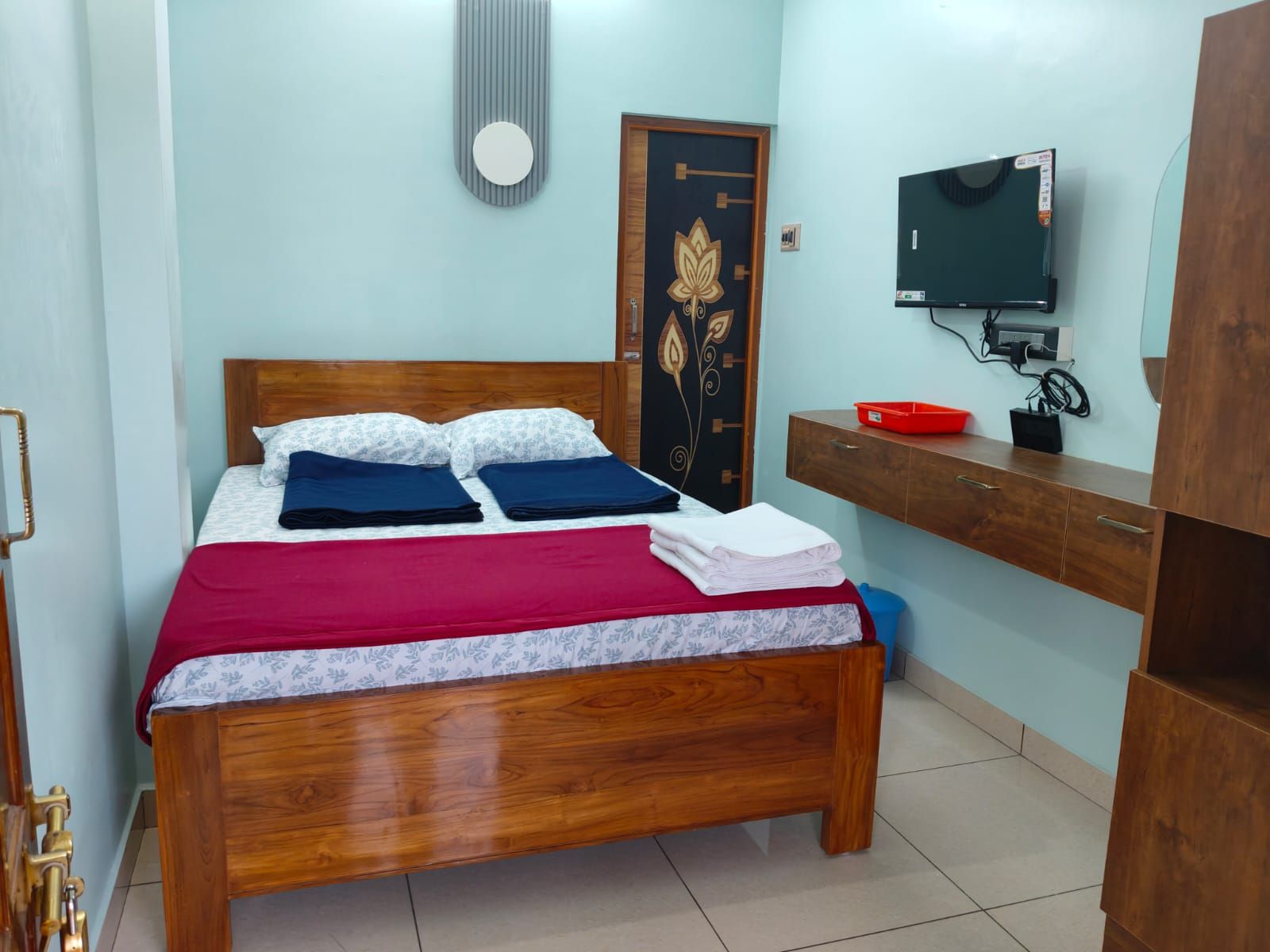 Un dormitorio con una cama de madera, un televisor sobre un mueble de madera, una puerta con diseño floral y una pequeña bandeja roja sobre un aparador de madera.