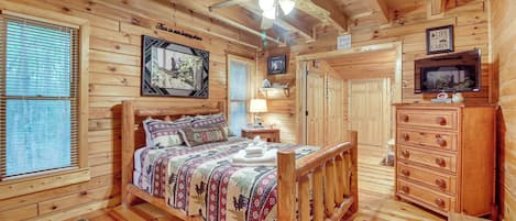 Un dormitorio de cabaña de madera con cama, cómoda, ventilador de techo y televisor.