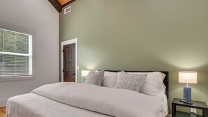 A bedroom with a large bed, two bedside lamps, a nightstand, and a window with blinds. - Luxe 4BR Cabin at Innsbruck Resort in Helen (Helen)