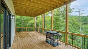 A covered outdoor deck with a grill, surrounded by a wooden railing, overlooking a forested area. - Luxe 4BR Cabin at Innsbruck Resort in Helen (Helen)