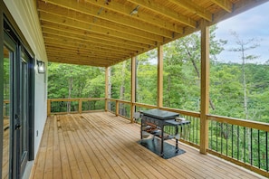 Eine überdachte Terrasse im Freien mit Grill, umgeben von einem Holzgeländer, mit Blick auf einen bewaldeten Bereich.