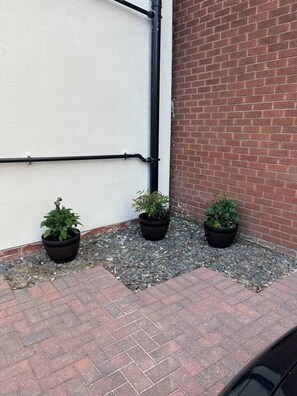 A corner of a building with a white wall, a brick wall, and three potted plants on a gravel and brick ground.