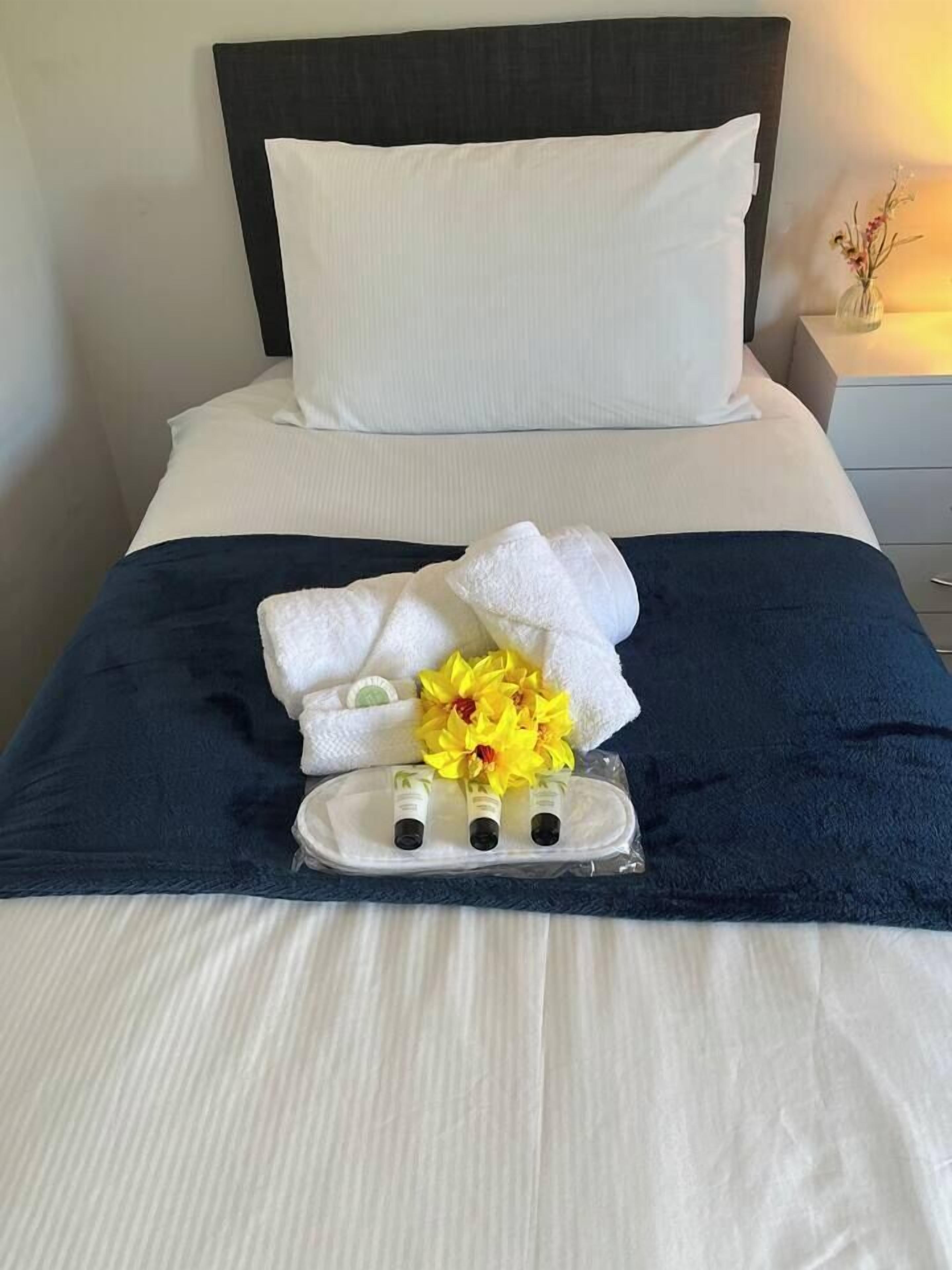 Een netjes opgemaakt bed met witte lakens, een donkerblauwe plaid en een dienblad met toiletartikelen en een bloemstuk.