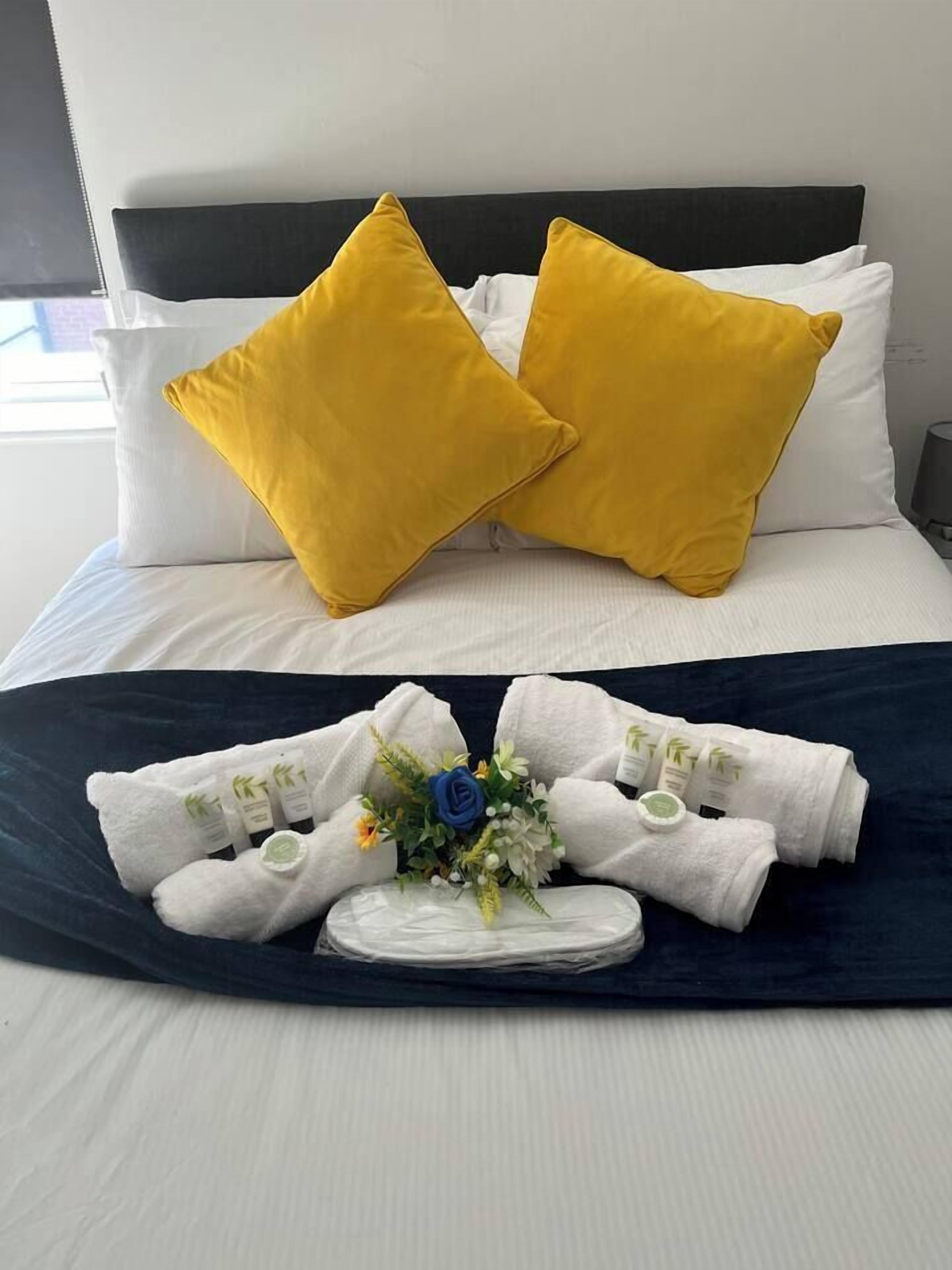 A bed with white and yellow pillows, a dark blue bedspread, and a tray with towels and toiletries.