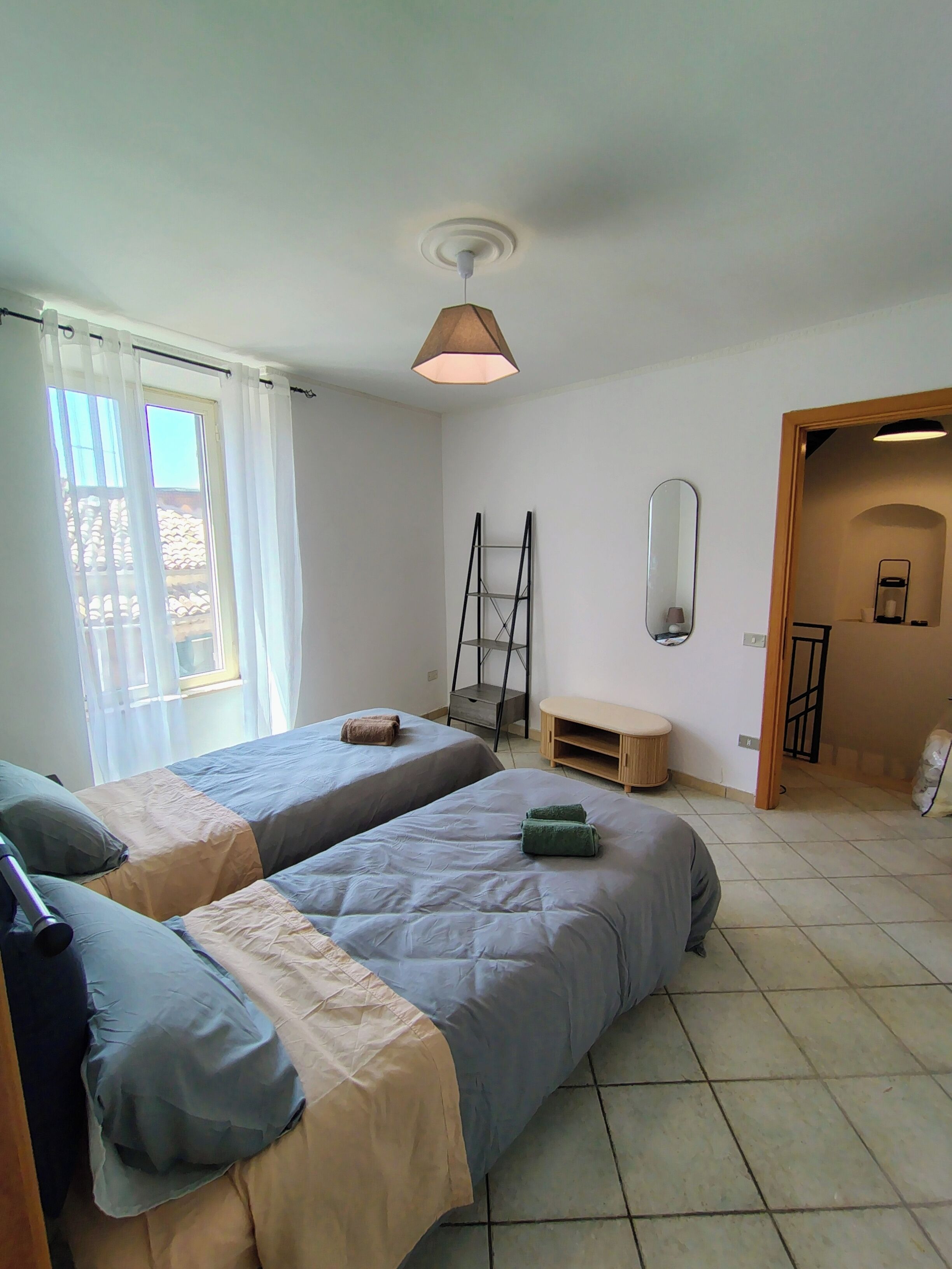 A bedroom with two beds, a ceiling fan, a window with curtains, a wooden bench, and a mirror.